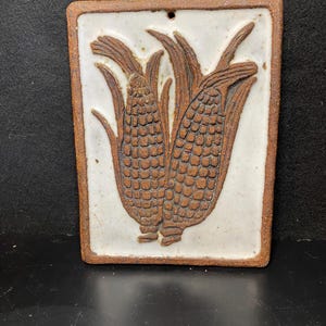 May include: A rectangular ceramic wall hanging featuring a relief of two ears of corn. The corn is brown with detailed kernels and green husks, set against a white background. The border is a darker brown.