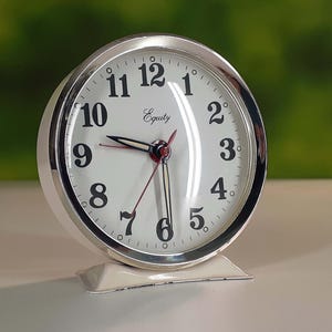 May include: A white and silver analog alarm clock with the brand name "Equity" on the face. The clock has black numerals, black hands, and a red second hand. The clock is approximately 4 inches tall.