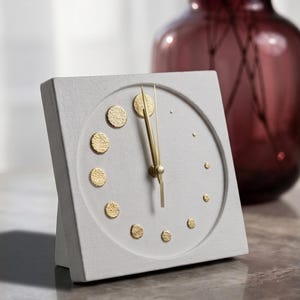 Può includere: Un orologio da scrivania quadrato in cemento grigio chiaro con un quadrante circolare incassato. L'orologio presenta lancette color oro e dodici indicatori circolari color oro di varie dimensioni che segnano le ore, con una finitura testurizzata.