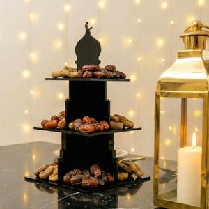 May include: A black, three-tiered serving stand with a mosque-shaped top, filled with dates and dried figs. A gold lantern with a lit candle sits to the right. The stand is on a black marble surface, with a string of lights in the background.