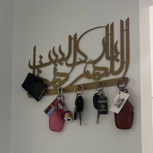 K&ouml;nnte beinhalten: Ein goldfarbener, wandmontierter Schl&uuml;sselhalter mit arabischer Kalligraphie. Der Halter hat mehrere Haken zum Aufh&auml;ngen von Schl&uuml;sseln, und an den Haken h&auml;ngen mehrere Schl&uuml;ssel und eine kleine schwarze Brieftasche.