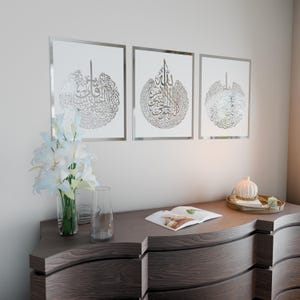 May include: Three silver framed wall art pieces with Arabic calligraphy in a white background. The art pieces are hanging on a white wall above a dark brown dresser.