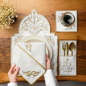 Op de afbeelding: Een wit geborduurd placemat, servet en bestekhouder met gouden en zilveren accenten. De set bevat een bord, een glas en goudkleurig bestek. De items zijn gerangschikt op een houten tafel.