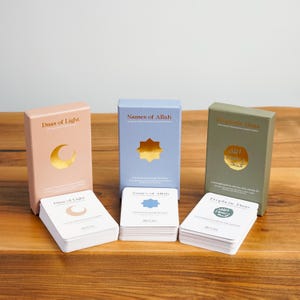 K&ouml;nnte beinhalten: Drei Kartensets mit religi&ouml;sen Themen. Die Boxen sind in Pastellrosa, Hellblau und Olivgr&uuml;n gehalten. Jede Box hat goldfarbene Akzente und Text. Die Karten sind wei&szlig; mit passenden goldenen oder blauen Akzenten. Auf einer Holzoberfl&auml;che.