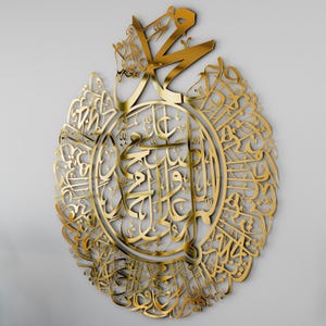 May include: A gold-colored metal wall art piece featuring intricate Arabic calligraphy in a circular design. The calligraphy is in a traditional style and is surrounded by a decorative border.