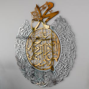 May include: A gold and silver metal wall art featuring intricate Arabic calligraphy. The design is circular with a central gold section and a silver outer ring. The calligraphy is in a traditional style and is likely a religious inscription.