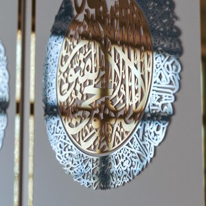 May include: A gold-colored metal wall decor with intricate Arabic calligraphy in a circular design. The calligraphy is surrounded by a silver-colored metal frame.