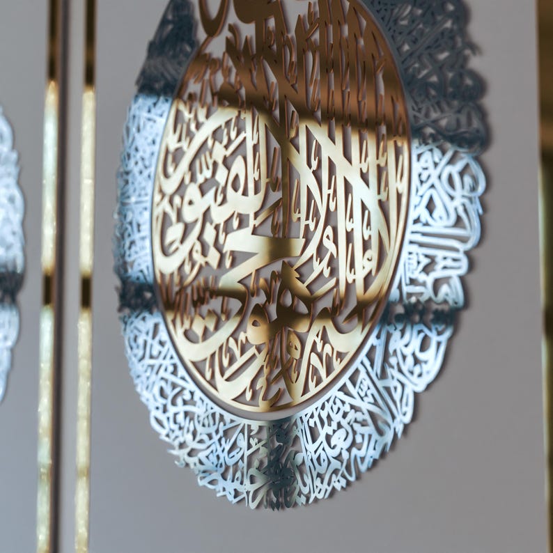 May include: A gold-colored metal wall decor with intricate Arabic calligraphy in a circular design. The calligraphy is surrounded by a silver-colored metal frame.