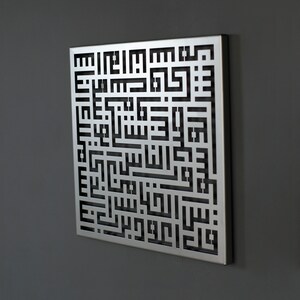 May include: A silver metal wall art piece with a black cutout design of Arabic calligraphy. The design is intricate and detailed, and the piece is a beautiful example of Islamic art.