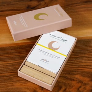 K&ouml;nnte beinhalten: Ein rosafarbener Kartensatz mit dem Titel "Duas of Light". Die Karten sind wei&szlig; mit einem Halbmond-Design und einem gelben Band. Die Box enth&auml;lt auch zwei Holzbl&ouml;cke. Der Text auf der Box lautet "Ein spiritueller Begleiter f&uuml;r Ihren Alltag."