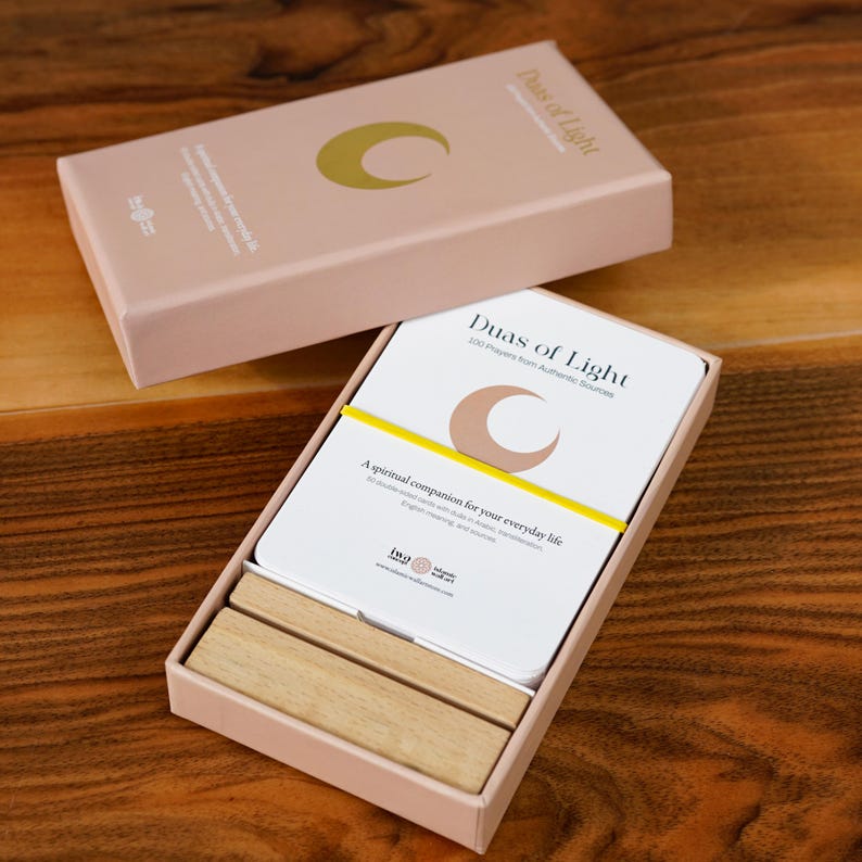 K&ouml;nnte beinhalten: Ein rosafarbener Kartensatz mit dem Titel "Duas of Light". Die Karten sind wei&szlig; mit einem Halbmond-Design und einem gelben Band. Die Box enth&auml;lt auch zwei Holzbl&ouml;cke. Der Text auf der Box lautet "Ein spiritueller Begleiter f&uuml;r Ihren Alltag."