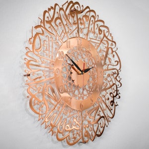 May include: A copper-colored wall clock with Arabic calligraphy around the clock face. The clock face has black hands and Roman numerals.