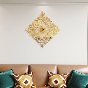 May include: A gold-colored, diamond-shaped wall clock with Arabic calligraphy. The clock features a sunburst design at the center and black clock hands. The clock is mounted on a white wall above a brown sofa with decorative pillows.