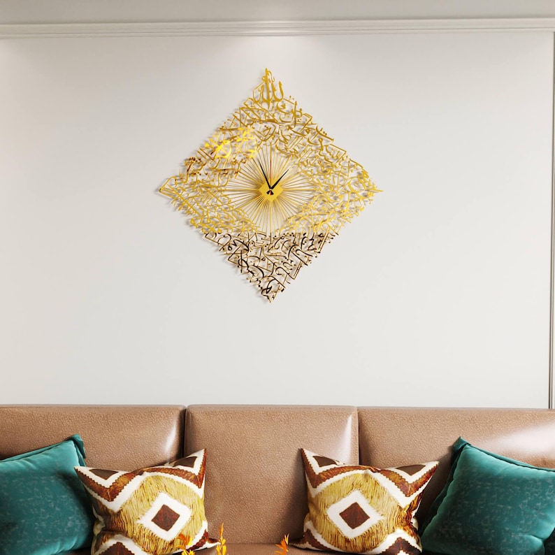 May include: A gold-colored, diamond-shaped wall clock with Arabic calligraphy. The clock features a sunburst design at the center and black clock hands. The clock is mounted on a white wall above a brown sofa with decorative pillows.
