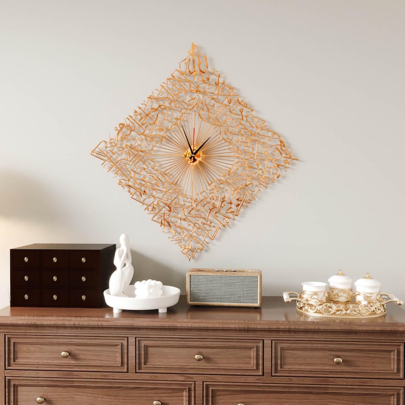May include: A gold-colored, diamond-shaped wall clock with Arabic calligraphy. The clock has a sunburst design in the center and black clock hands. The clock is mounted on a light gray wall above a wooden dresser with decorative items.