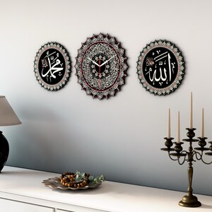 Op de afbeelding: Drie ronde wandklokken met zwarte frames en ingewikkelde witte ontwerpen. De middelste klok heeft een witte wijzerplaat met zwarte wijzers en Arabische cijfers. De andere twee klokken tonen Arabische kalligrafie in wit op een zwarte achtergrond.