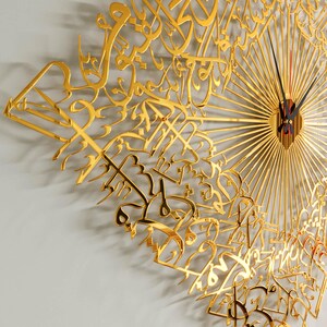 May include: A gold-colored wall clock with intricate Arabic calligraphy design. The clock features a sunburst pattern and black clock hands with a red second hand. The clock's design is a decorative piece for home decor.