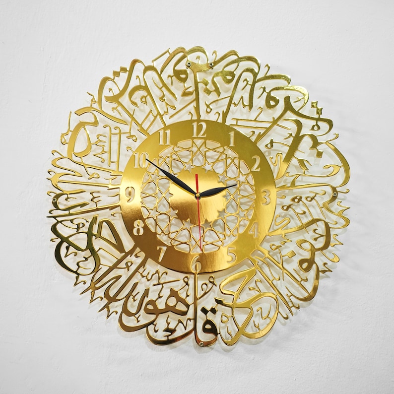 May include: Gold-colored wall clock with Arabic calligraphy surrounding the clock face. The clock face has black hands and Roman numerals.