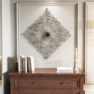 May include: A silver metal wall clock with Arabic calligraphy in a diamond shape. The clock has black hands and is mounted on a light gray wall. The clock is above a dark brown wooden dresser with books and a candle.