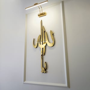 May include: A gold-colored Arabic calligraphy art piece mounted on a white wall, illuminated by a gold-toned picture light. The calligraphy is in a stylized script, and the wall has a decorative frame.