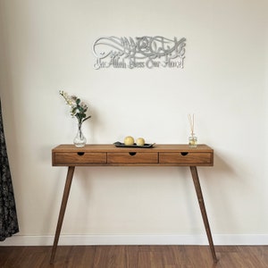 May include: A silver metal wall decor with Arabic calligraphy that reads "May Allah Bless Our Home". The decor is hanging on a white wall above a wooden console table with three drawers. The table has a light brown finish and is sitting on a hardwood floor.