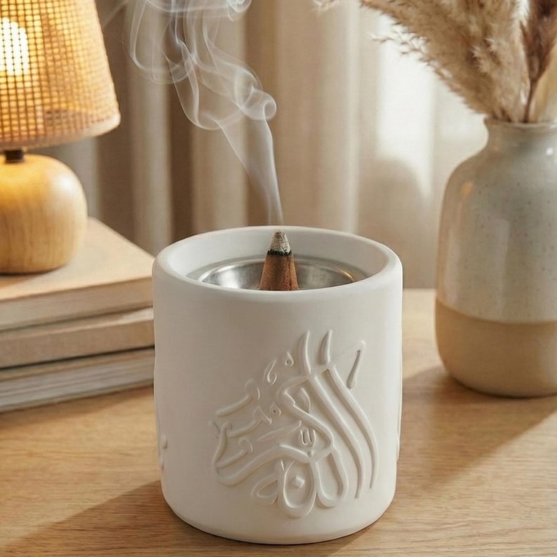 Peut inclure: Un porte-encens en c&eacute;ramique blanche avec un insert en m&eacute;tal argent&eacute;, d&eacute;gageant des volutes de fum&eacute;e. Le support pr&eacute;sente un motif en relief de calligraphie arabe. Un c&ocirc;ne d'encens allum&eacute; est &agrave; l'int&eacute;rieur.