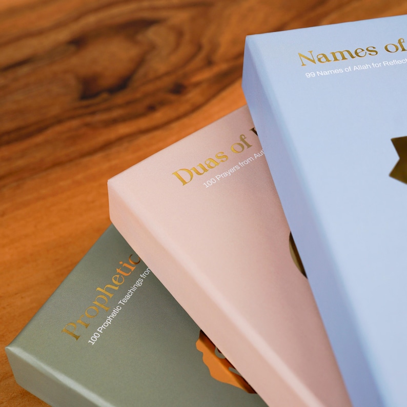 K&ouml;nnte beinhalten: Drei Hardcover-B&uuml;cher in Pastellfarben: Hellblau, Rosa und Gr&uuml;n. Goldene Schrift auf den Einb&auml;nden lautet "Names of", "Duas of" und "Prophetic". Die B&uuml;cher sind auf einer Holzoberfl&auml;che gestapelt.