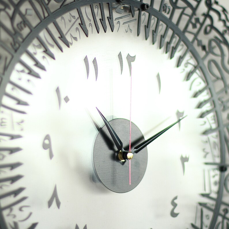 May include: A black and white clock with Arabic numerals. The clock face is white with black numbers and hands. The clock is framed in black metal with Arabic script.