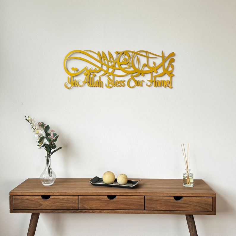 May include: Gold metal wall art with Arabic calligraphy and the text "Ya Allah Bless Our Home". The art is hanging on a white wall above a wooden table with two decorative spheres and a diffuser.
