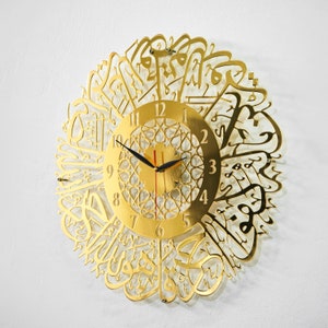 May include: Gold-colored wall clock with Arabic calligraphy surrounding the clock face. The clock face has a gold background with black hands and black Roman numeral markings.