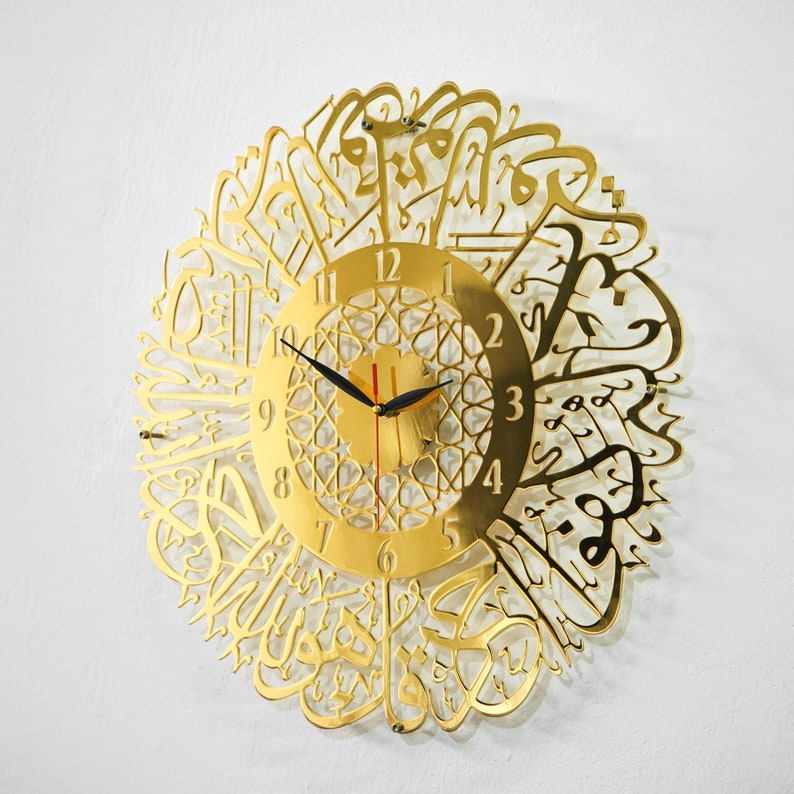 May include: Gold-colored wall clock with Arabic calligraphy surrounding the clock face. The clock face has a gold background with black hands and black Roman numeral markings.
