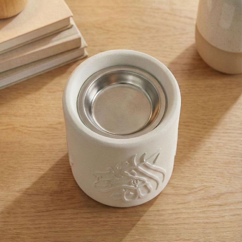 Peut inclure: Un chauffe-huile en c&eacute;ramique cr&egrave;me avec un insert en m&eacute;tal argent&eacute;. Le chauffe-huile pr&eacute;sente un motif floral en relief sur le c&ocirc;t&eacute;. Une pile de livres et une tasse en c&eacute;ramique sont en arri&egrave;re-plan.