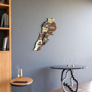 May include: A wooden map of Lebanon with the names of each region written in Arabic. The map is mounted on a grey wall with a glass-topped table and a silver metal stand in the foreground.