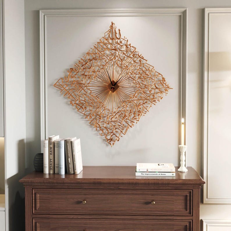 May include: A gold-colored, diamond-shaped wall clock with Arabic calligraphy. The clock has a sunburst design around the center and is mounted on a light gray wall. The clock is above a dark brown wooden dresser.