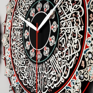 Op de afbeelding: Een zwarte klok met Arabische kalligrafie rond de wijzerplaat. De klok heeft witte en rode wijzers en een decoratieve rand met rode, witte en blauwe accenten.