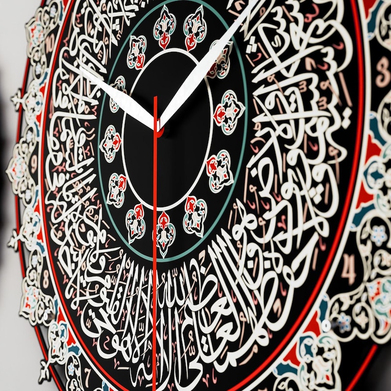 Op de afbeelding: Een zwarte klok met Arabische kalligrafie rond de wijzerplaat. De klok heeft witte en rode wijzers en een decoratieve rand met rode, witte en blauwe accenten.