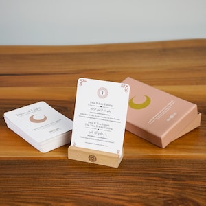K&ouml;nnte beinhalten: Ein Satz wei&szlig;er Karten mit Text und einem Halbmond-Logo, pr&auml;sentiert mit einem Kartenhalter aus Holz und einer rosa Schachtel. Die Karten tragen die Titel "Duas of Light" und "Dua Before Eating."