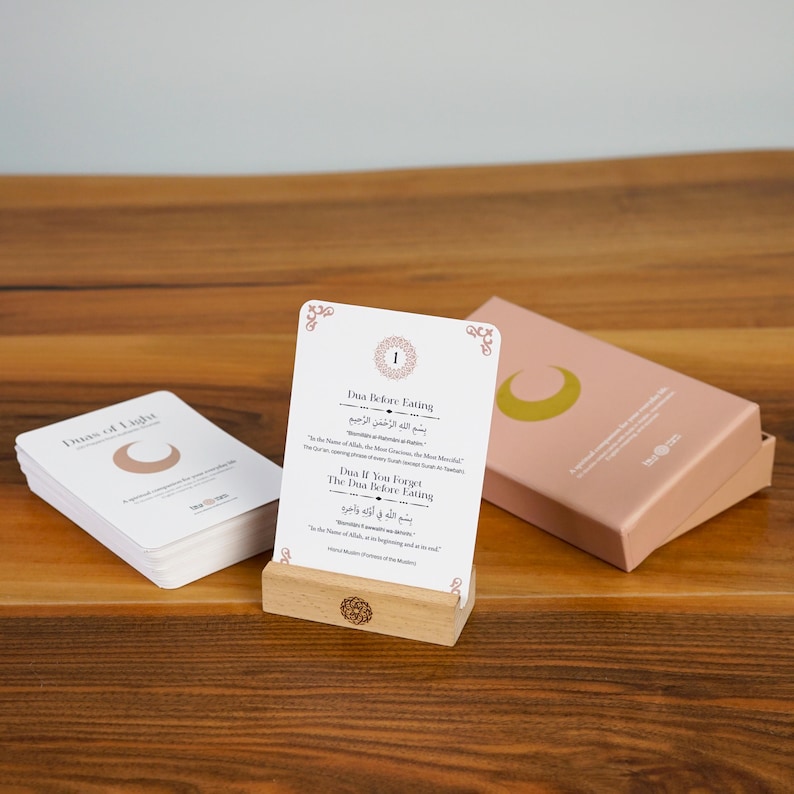K&ouml;nnte beinhalten: Ein Satz wei&szlig;er Karten mit Text und einem Halbmond-Logo, pr&auml;sentiert mit einem Kartenhalter aus Holz und einer rosa Schachtel. Die Karten tragen die Titel "Duas of Light" und "Dua Before Eating."