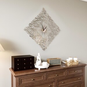 May include: A decorative wall clock with a diamond-shaped design, featuring intricate metallic script and radial spokes. The clock is mounted above a wooden dresser with a black jewelry box, a white sculpture, and a gold-trimmed tray.