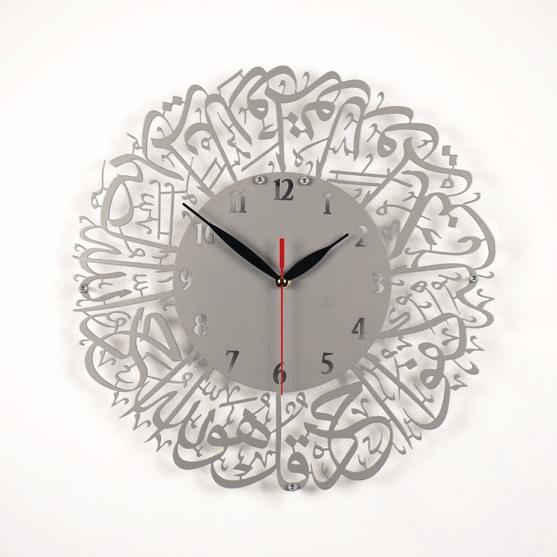 K&ouml;nnte beinhalten: Eine graue Wanduhr aus Metall mit arabischer Kalligraphie um das Zifferblatt. Die Uhr hat schwarze Zeiger und einen roten Sekundenzeiger. Die Zahlen auf dem Zifferblatt sind schwarz.