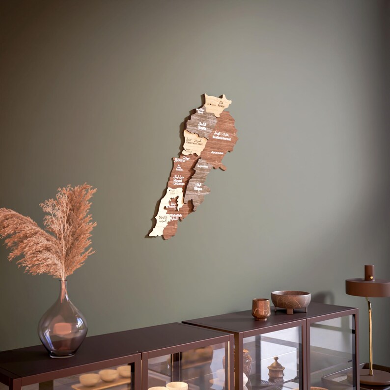 May include: A wooden map of Lebanon with the names of each region in Arabic and English. The map is mounted on a green wall and is surrounded by a brown wooden shelf with glass doors.