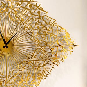 May include: A gold-colored wall clock with a unique design. The clock features a sunburst pattern with radiating lines and intricate Arabic calligraphy. The clock hands are black, and the clock face is square. The clock is mounted on a white wall.