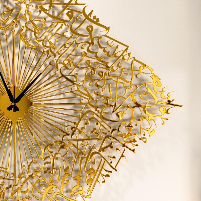 May include: A gold-colored wall clock with a unique design. The clock features a sunburst pattern with radiating lines and intricate Arabic calligraphy. The clock hands are black, and the clock face is square. The clock is mounted on a white wall.