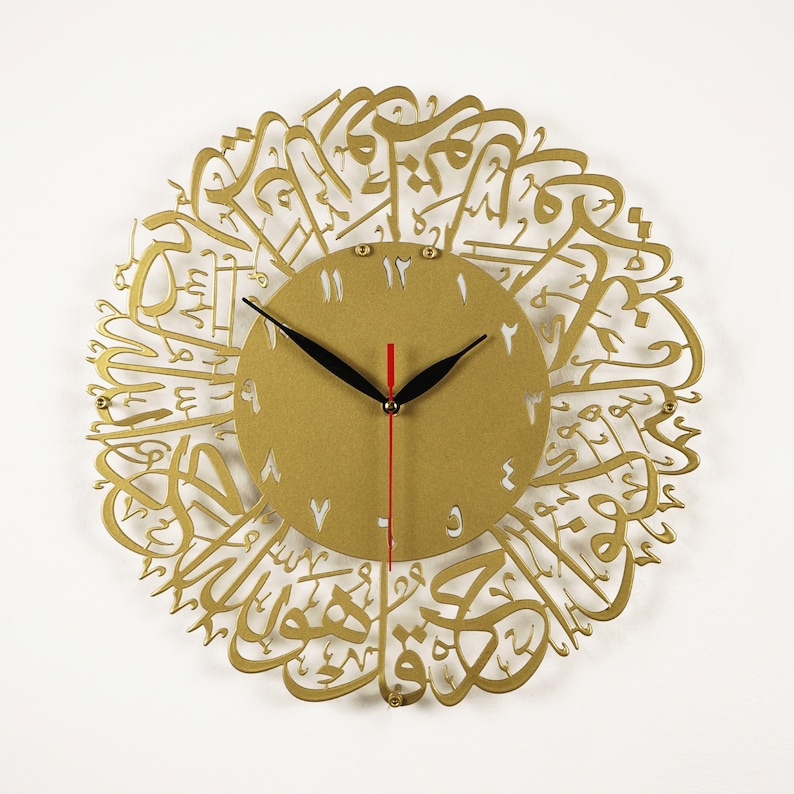 K&ouml;nnte beinhalten: Goldfarbene Wanduhr mit arabischer Kalligraphie um das Zifferblatt. Das Zifferblatt hat schwarze Zeiger und wei&szlig;e r&ouml;mische Ziffern.
