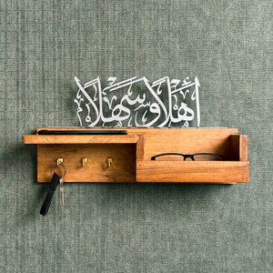 Op de afbeelding: Een houten sleutelhanger met een plank en een klein lade. De sleutelhanger heeft drie haken en een metalen bord met Arabische kalligrafie waarop "Alhamdulillah" staat.