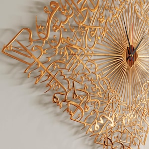 May include: Gold-colored wall clock featuring Arabic calligraphy. The clock has a sunburst design with radiating lines and a central clock face with black hands and a red second hand. The clock is mounted on a light gray wall.