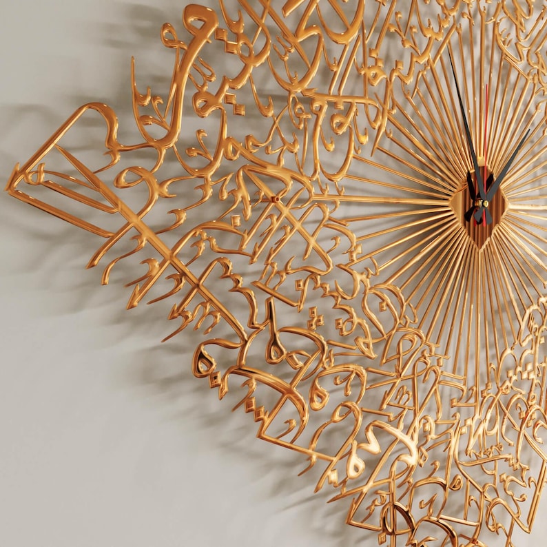 May include: Gold-colored wall clock featuring Arabic calligraphy. The clock has a sunburst design with radiating lines and a central clock face with black hands and a red second hand. The clock is mounted on a light gray wall.