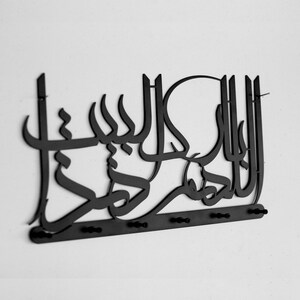K&ouml;nnte beinhalten: Schwarze Wanddekoration aus Metall mit arabischer Kalligraphie. Das Design beinhaltet einen Kreis und eine Reihe von Haken unten.