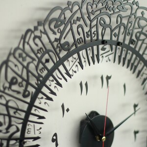 May include: A black metal clock with Arabic numerals and a black background. The clock face is surrounded by a decorative border of Arabic calligraphy.