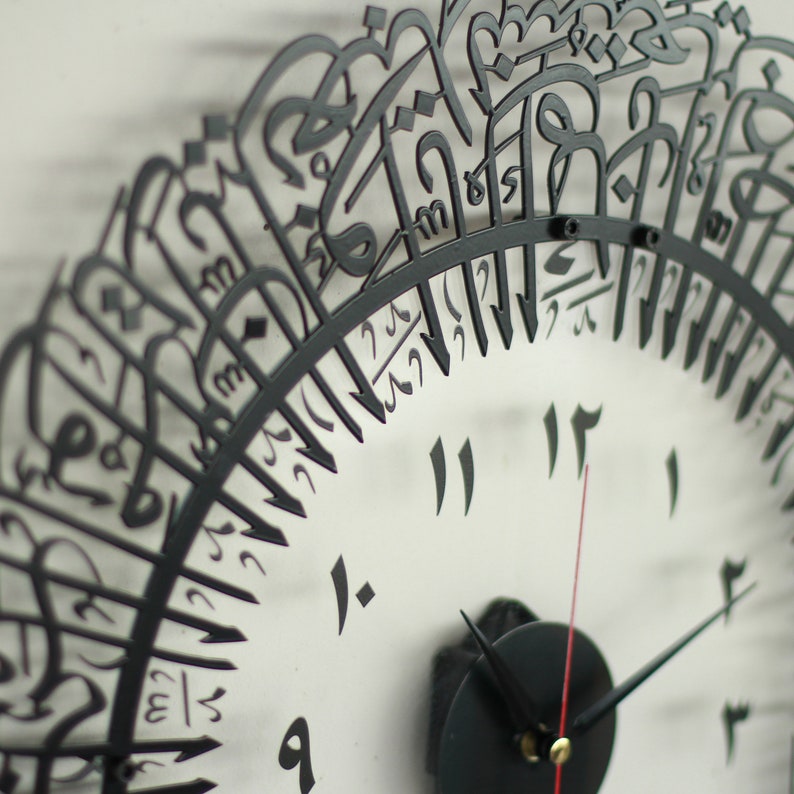 May include: A black metal clock with Arabic numerals and a black background. The clock face is surrounded by a decorative border of Arabic calligraphy.